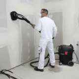 Operaio con maschera e occhiali usa una levigatrice con aspirazione per preparare il muro, usando la carta vetrata per una finitura perfetta.