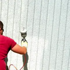 Uomo con maglietta rossa e occhiali da sole sta pitturando casa a spruzzo con una pistola.