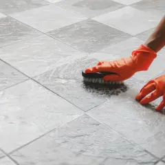 Mani con guanti arancioni puliscono un pavimento piastrellato con una spazzola, mostrando come pulire il pavimento dopo la posa.