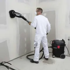 Operaio con maschera e occhiali usa una levigatrice con aspirazione per preparare il muro, usando la carta vetrata per una finitura perfetta.