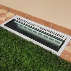 Bocche di lupo moderne con griglia metallica e illuminazione integrata, incassate tra prato verde e pavimentazione in cotto.