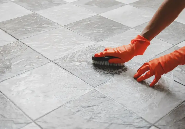 Mani con guanti arancioni puliscono un pavimento piastrellato con una spazzola, mostrando come pulire il pavimento dopo la posa.