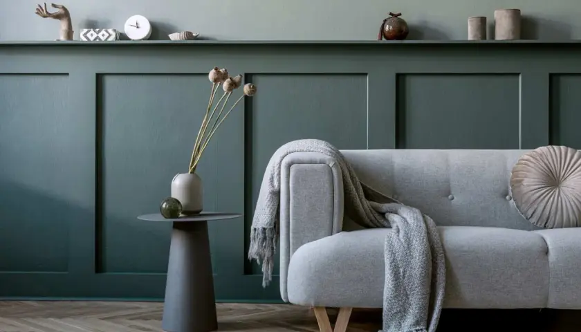 Soggiorno con divano grigio, coperta e decorazioni su una parete con effetto boiserie con pittura verde petrolio.