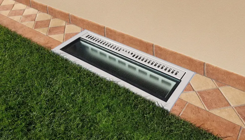 Bocche di lupo moderne con griglia metallica e illuminazione integrata, incassate tra prato verde e pavimentazione in cotto.
