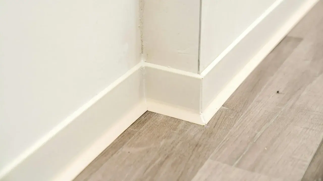 Angolo di un battiscopa in ceramica bianca, pronto per essere incollato su un pavimento in laminato grigio.