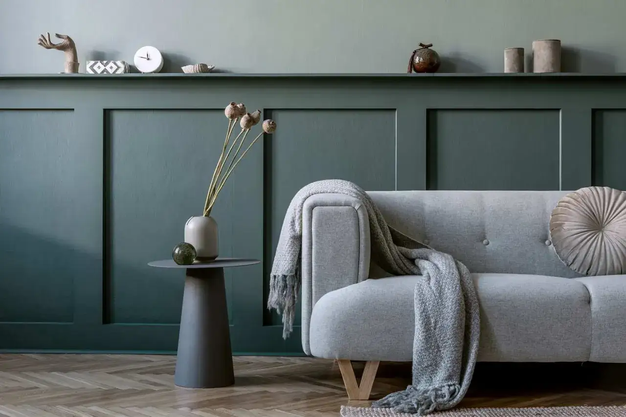 Un elegante divano grigio con coperta, accanto a un tavolino con vaso e fiori secchi, su una parete con boiserie fai da te.