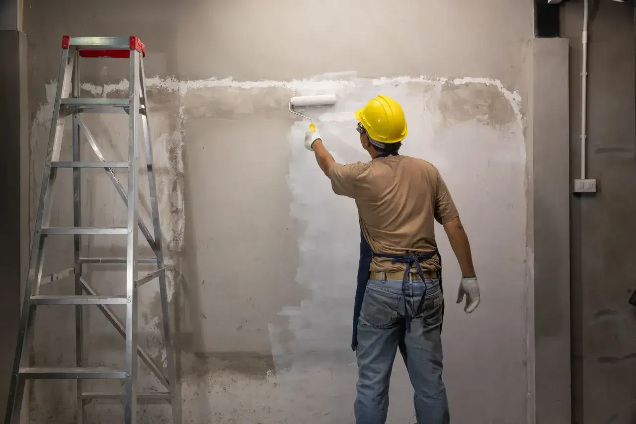 Operaio con casco giallo stende pittura bianca su muro grezzo. Vicino, una scala a pioli. Si prepara la superficie, forse il fissativo si può mischiare nella pittura.
