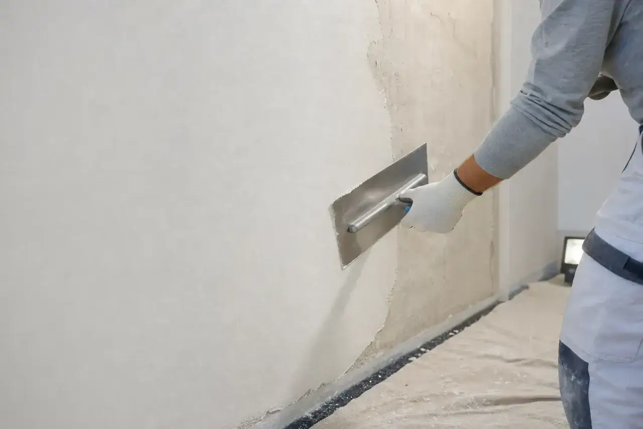 Un operaio con guanti bianchi esegue la rasatura di un muro con una spatola metallica, creando una superficie liscia e uniforme.